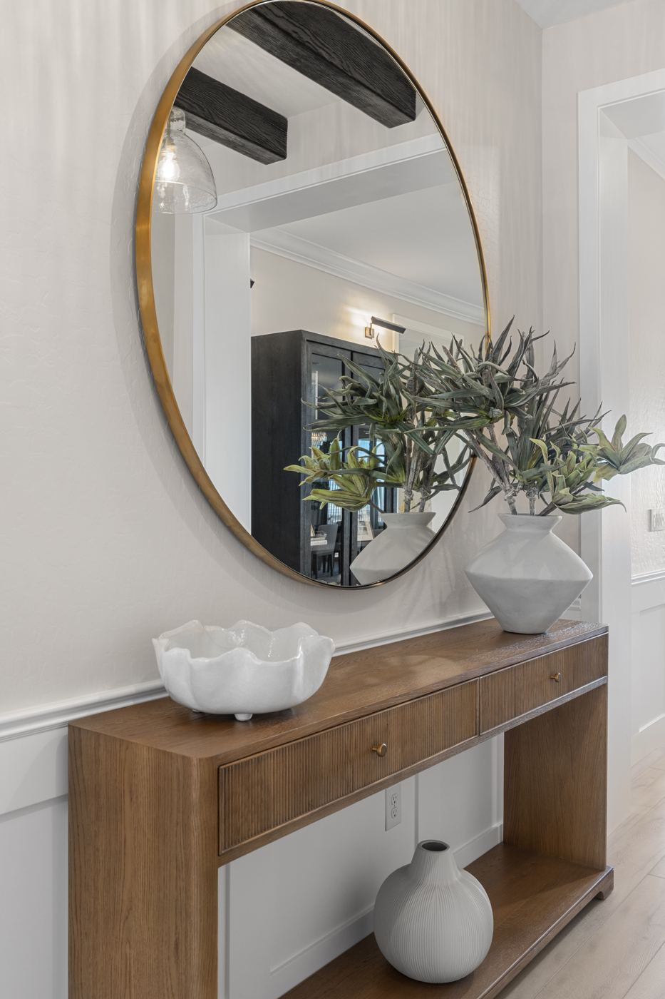 Oak console with a round brass mirror, a white scalloped bowl, and a sculptural vase of greenery