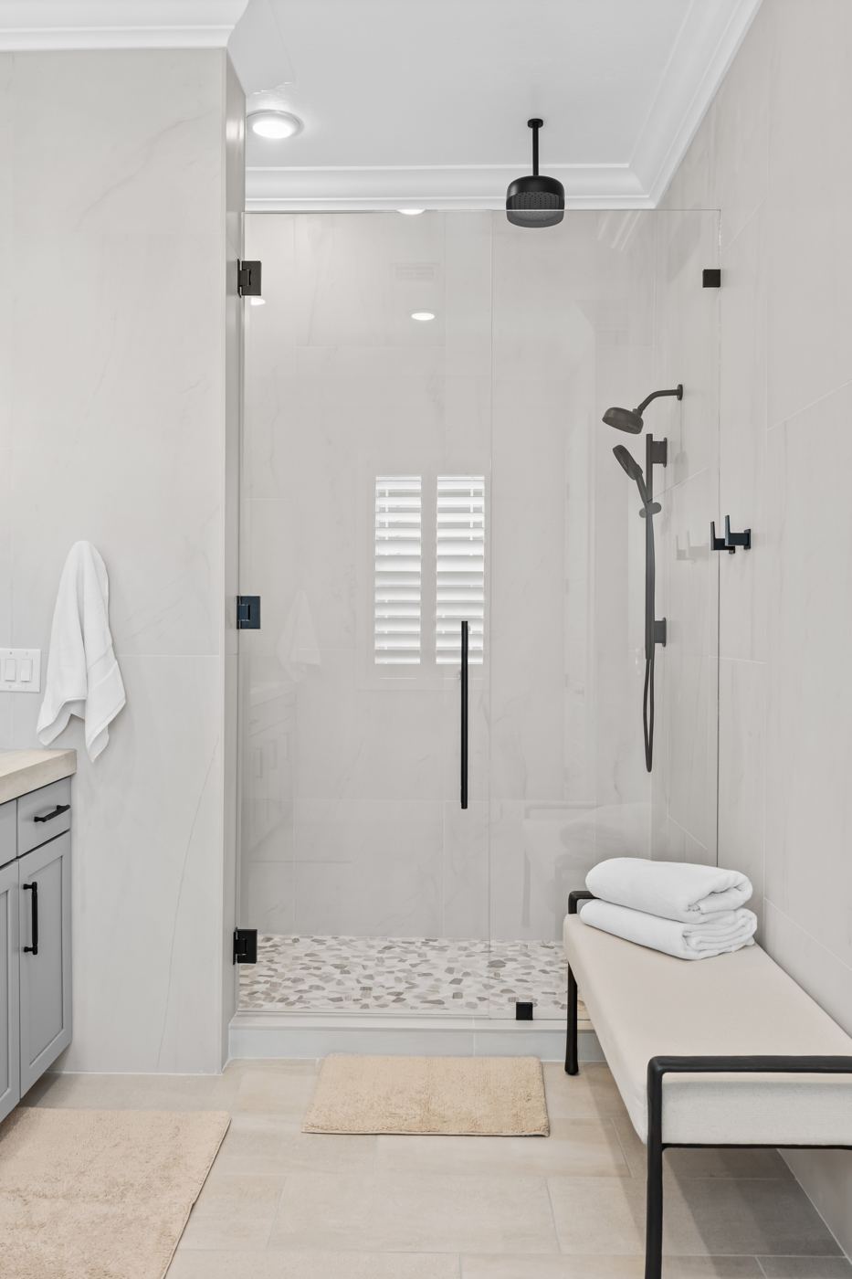 Walk-in shower with frameless glass, matte black fixtures, and a pebble floor, beside an upholstered bench